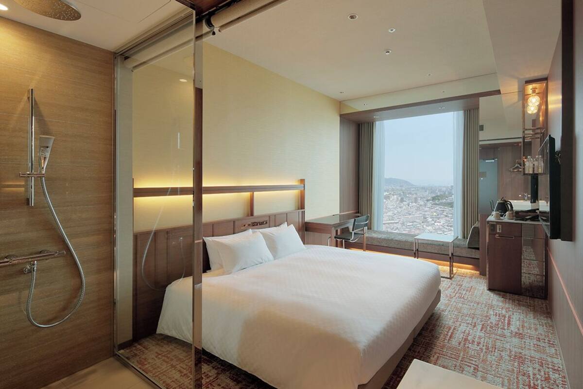 Een hotelkamer met een bed, douche, bureau, stoel, televisie en uitzicht op de stad.