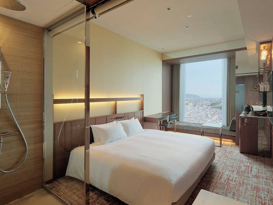 Habitación de hotel con cama, ducha, escritorio, silla, televisor y vistas de la ciudad.