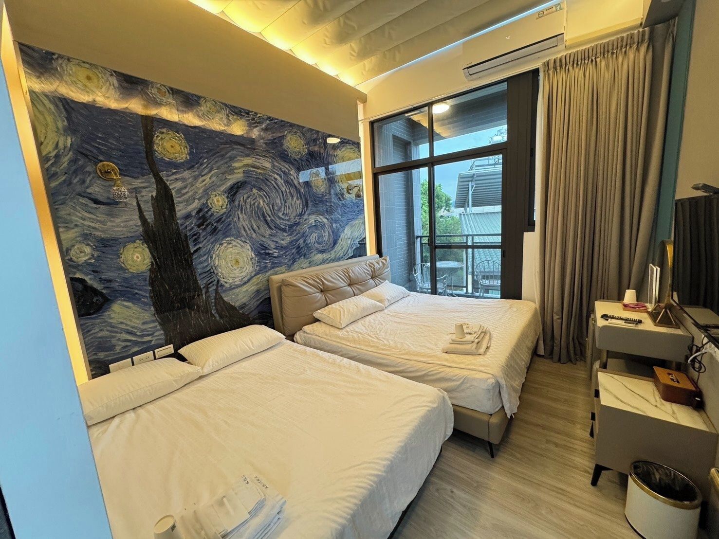 A hotel room with two beds, a large painting on the wall, a window with curtains, and a desk with a lamp and a trash can.
