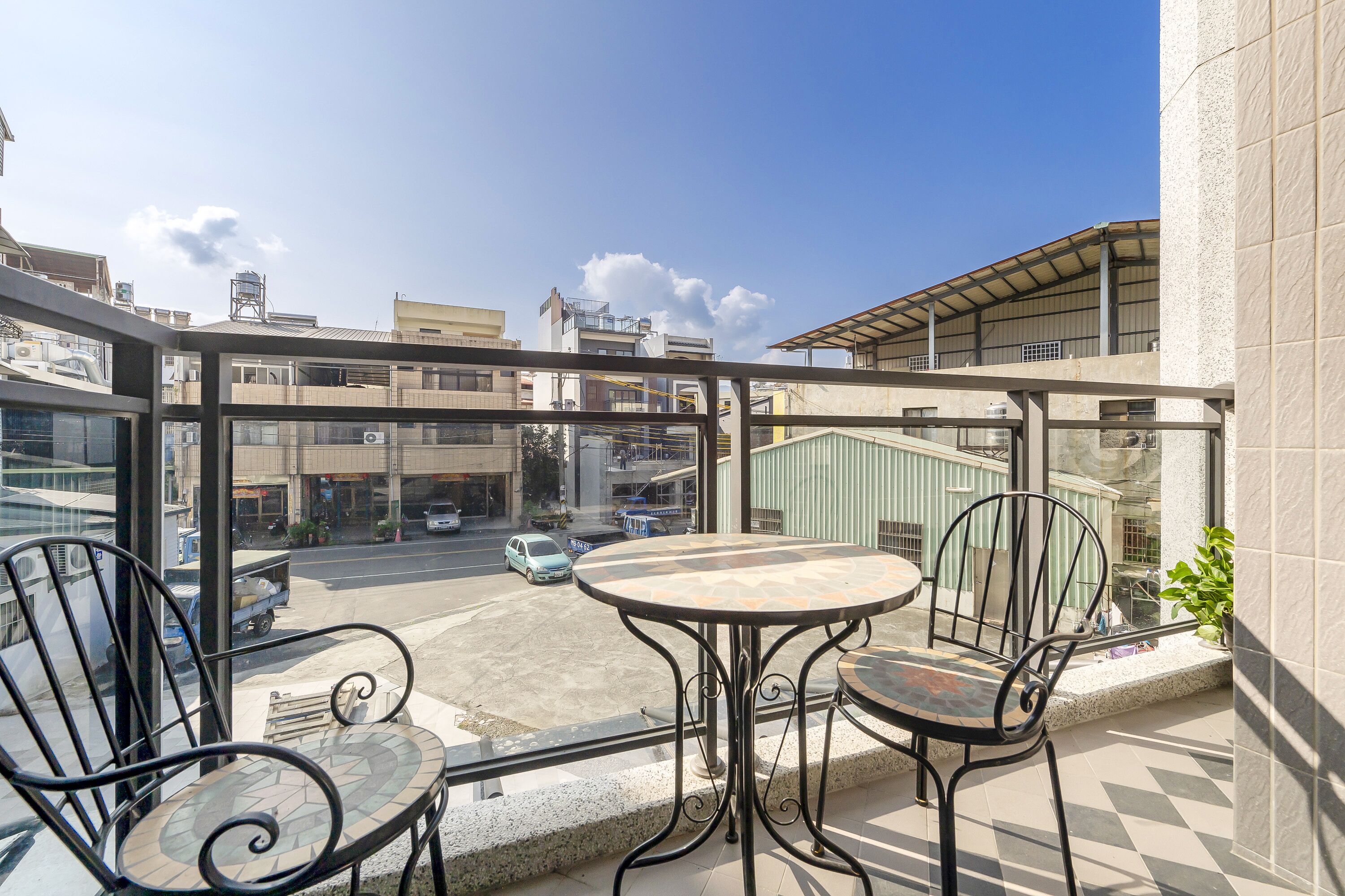 A balcony with a table and chairs, overlooking a street with buildings and parked cars.