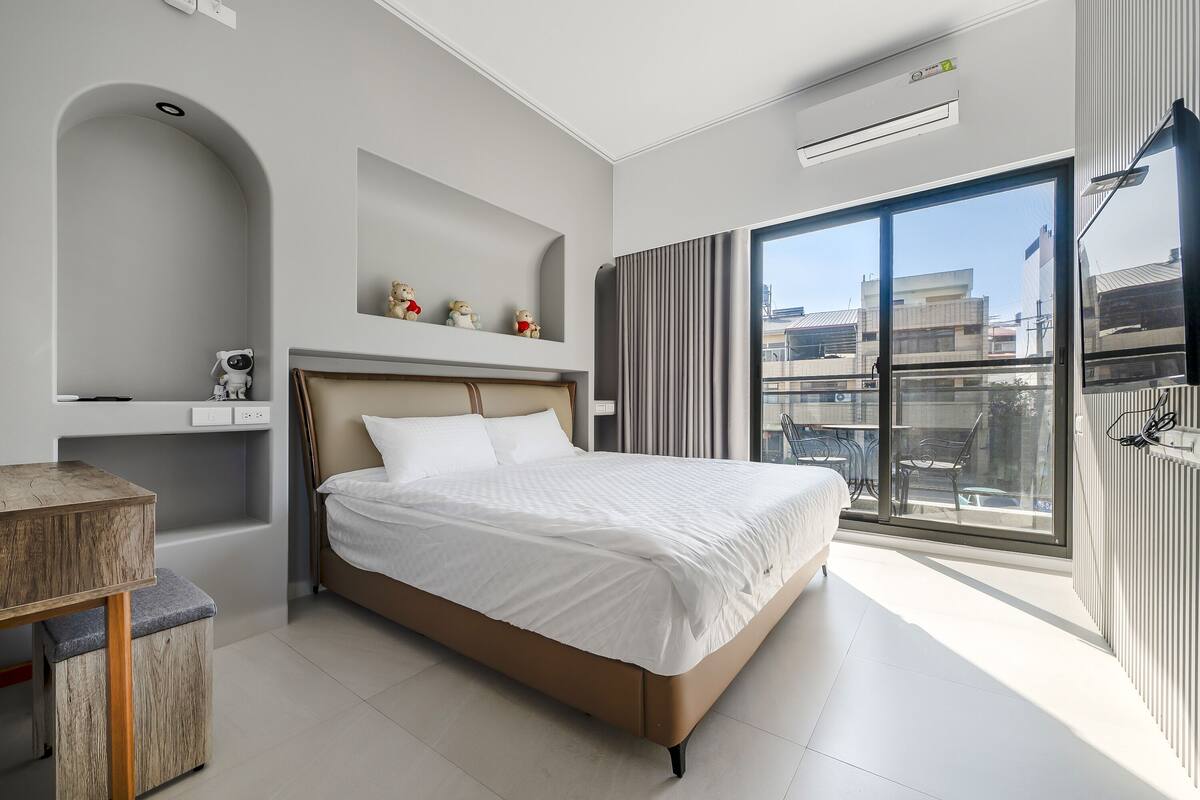 A modern hotel room with a large bed, a small desk, and a view of the city through large windows.