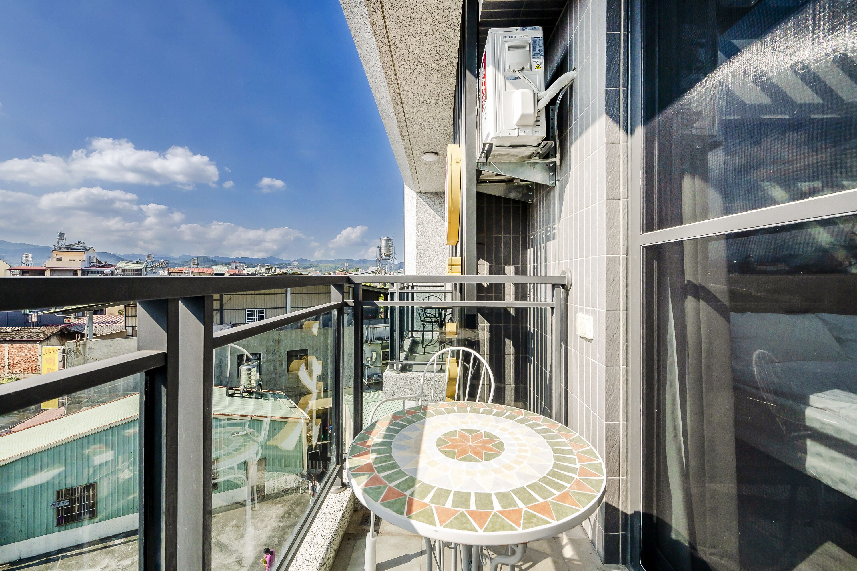 A balcony with a glass railing, a tiled table, and a view of buildings and mountains.