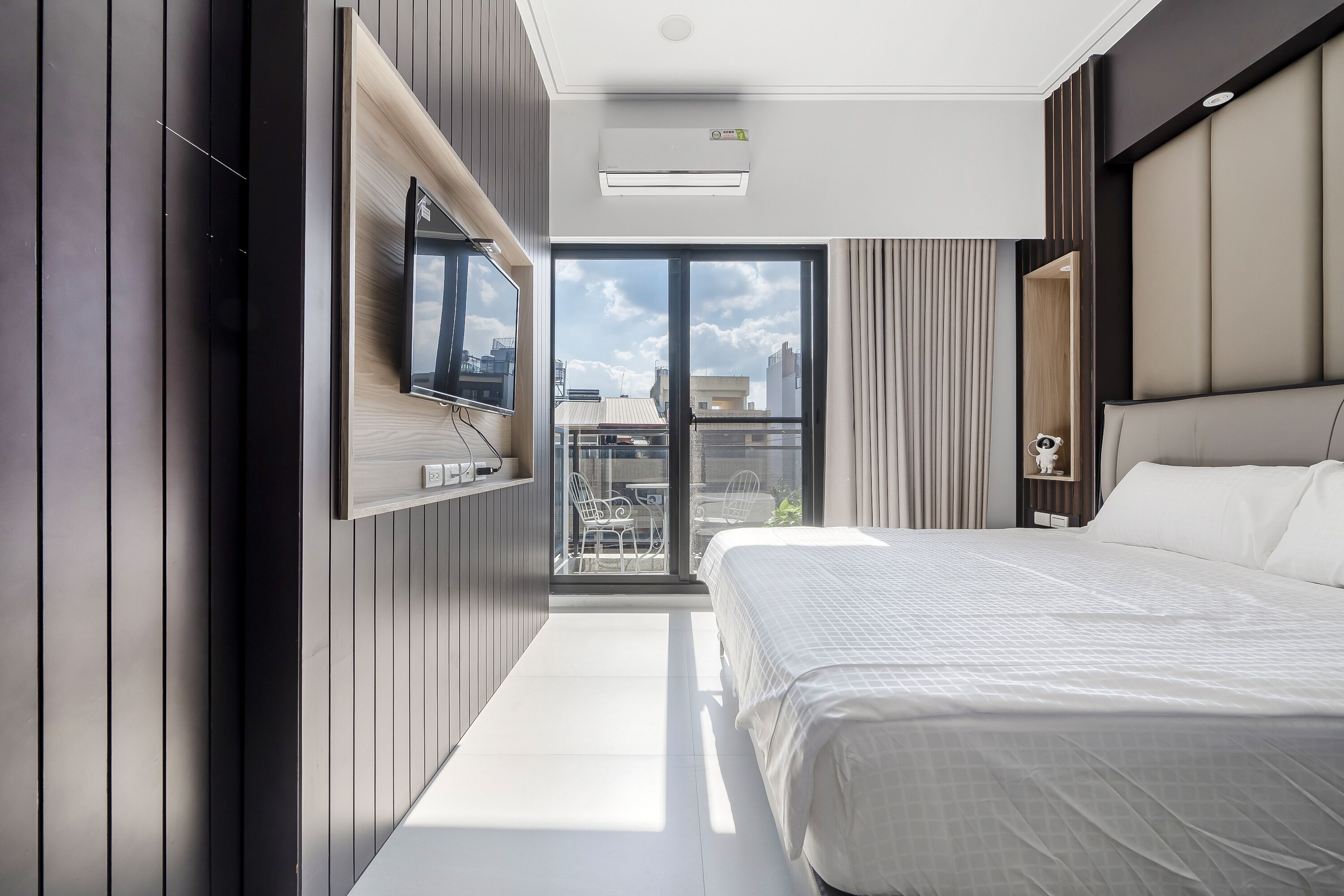 A modern hotel room with a large bed, a flat-screen TV mounted on the wall, and a balcony with a city view.