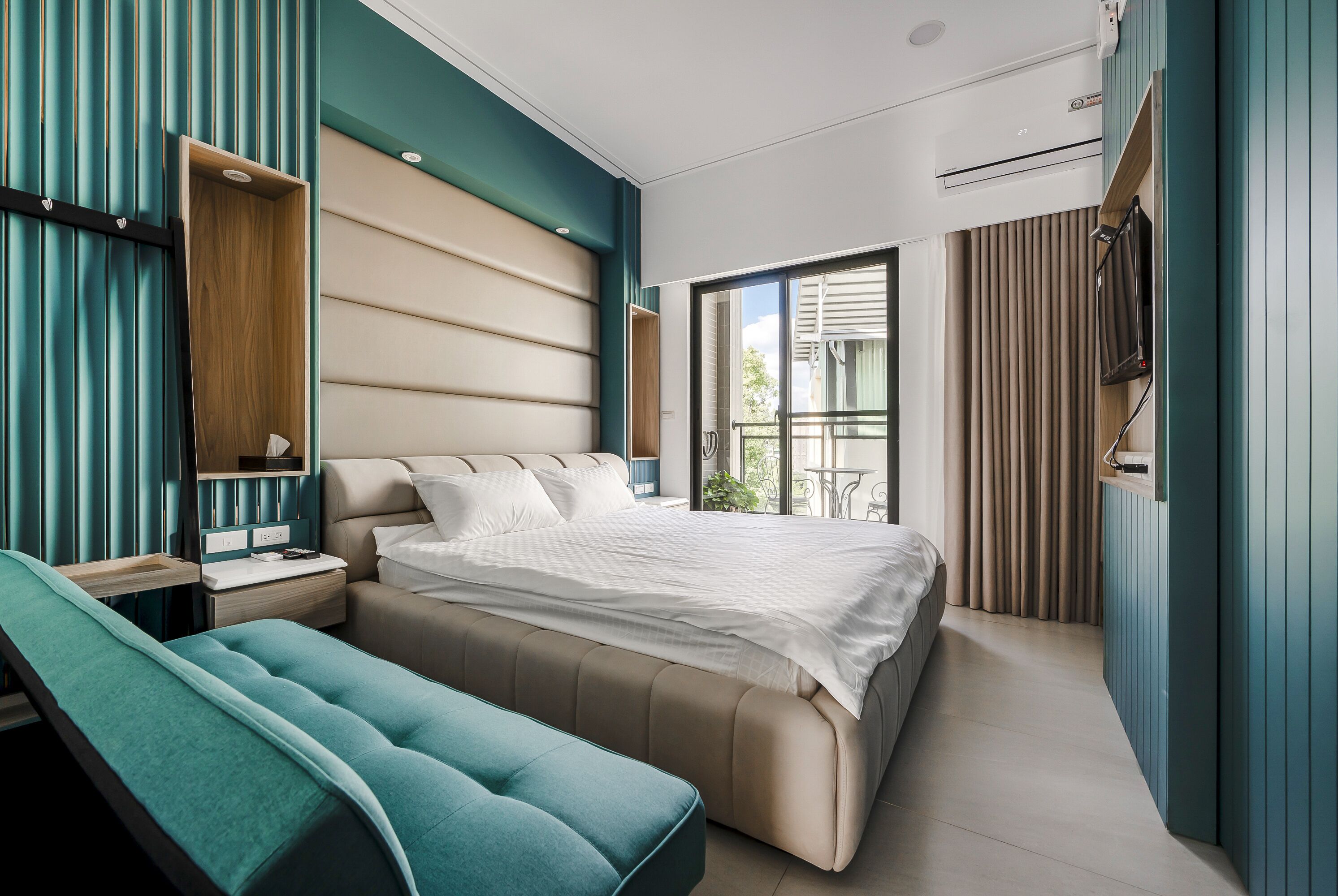 A modern bedroom with a large bed, a bench, a bedside table, and a view of the outdoors.