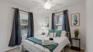 Ein Schlafzimmer mit Bett, Deckenventilator, Nachttisch, Lampe, einem gerahmten Kunstwerk mit Meeresschweine und einer Topfpflanze.