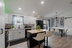 A modern kitchen with white cabinets, a central island with bar stools, a dining area with a table and chairs, and a large window with curtains. - Newly Renovated 4BR Beach Retreat: Walk to beach,Games, Pet Friendly, Near DT (St. Augustine)