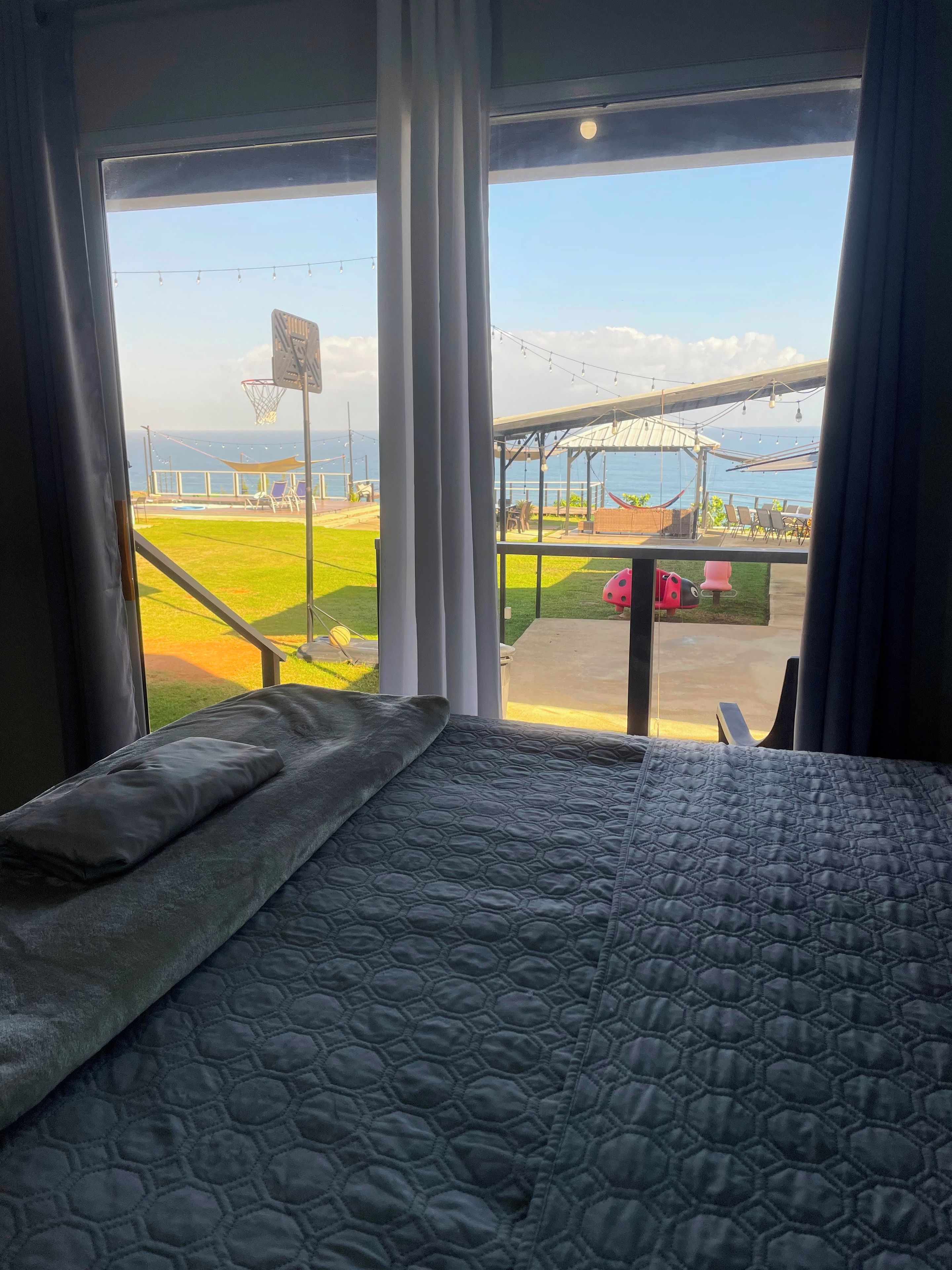 A bed with a quilted cover, a view of a sports court, and a building with a roof structure.