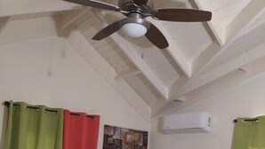 Ein Schlafzimmer mit einem Bett, einem Deckenventilator, grün-roten Vorhängen, einem kleinen Nachttisch und einer Klimaanlage.