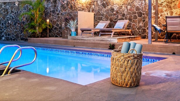 A pool area with lounge chairs, a wicker basket with towels, and a stone wall. - Luxurious Makai Villa with Pool and Ocean Views (Kailua Kona)