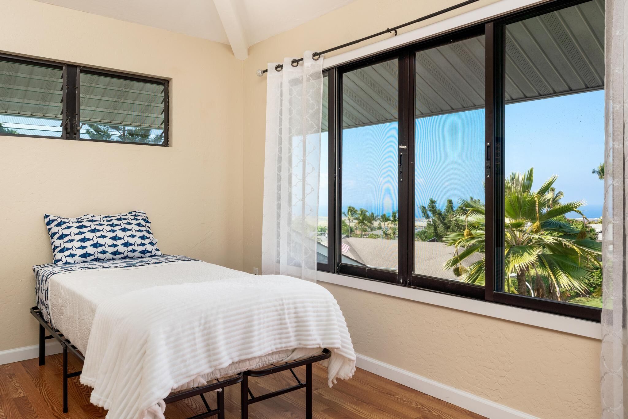 A bedroom with a bed, a window with a view of palm trees, and a curtain.