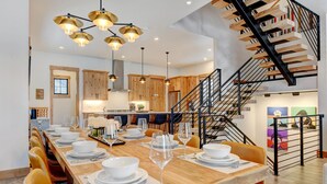 A modern kitchen with wooden cabinets, a dining area with a long table set for six, and a staircase with a wooden railing.