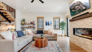 A spacious living room with a large sectional sofa, a wooden coffee table, and a fireplace. There are two windows with wooden frames and a large picture on the wall.
