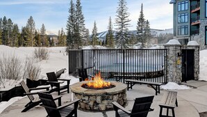 A stone fire pit surrounded by black chairs, with a snow-covered landscape and mountains in the background.