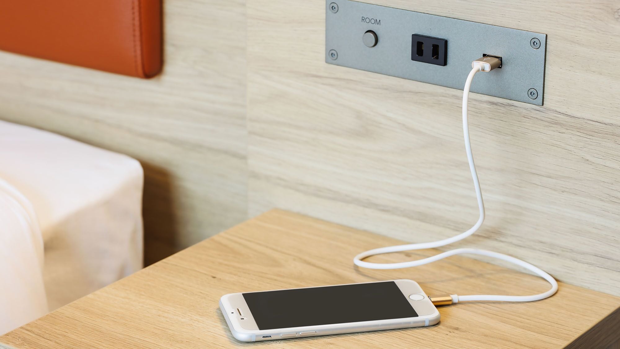 A smartphone connected to a power outlet on a wooden nightstand in a hotel room.