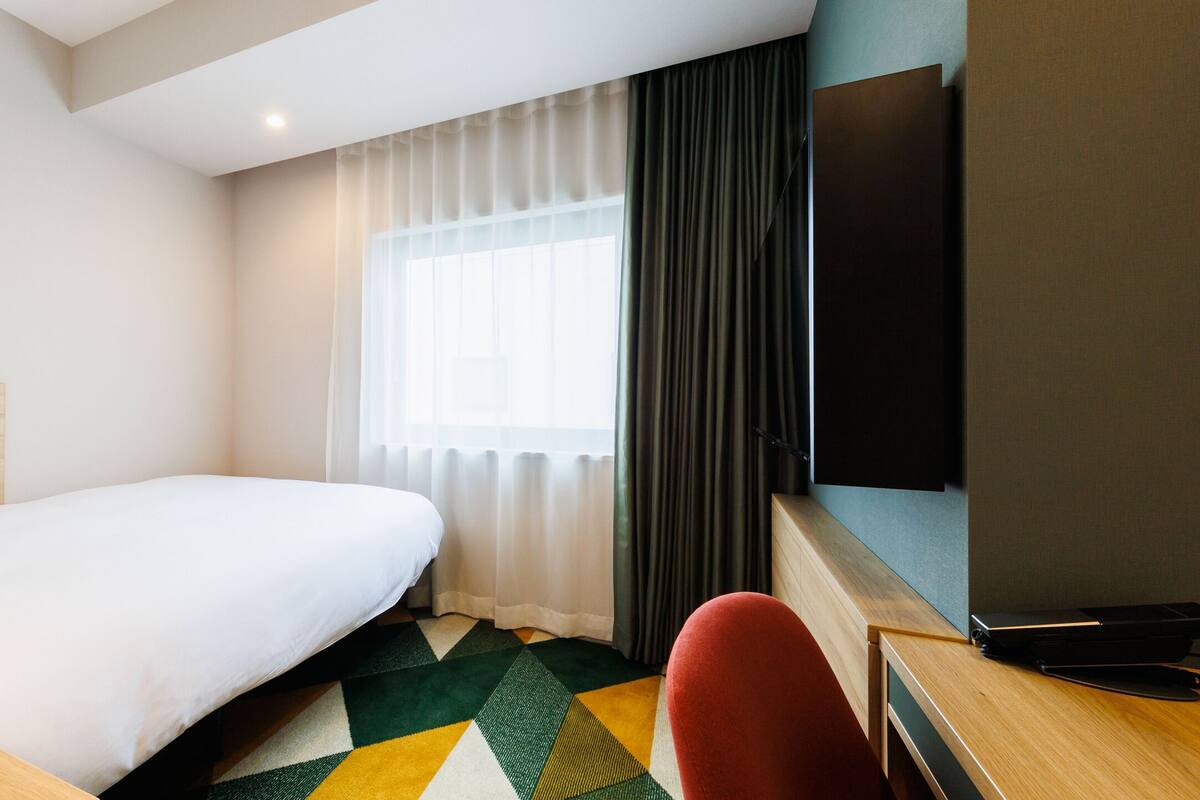 A hotel room with a bed, a desk with a flat-screen TV, a chair, and a colorful rug.