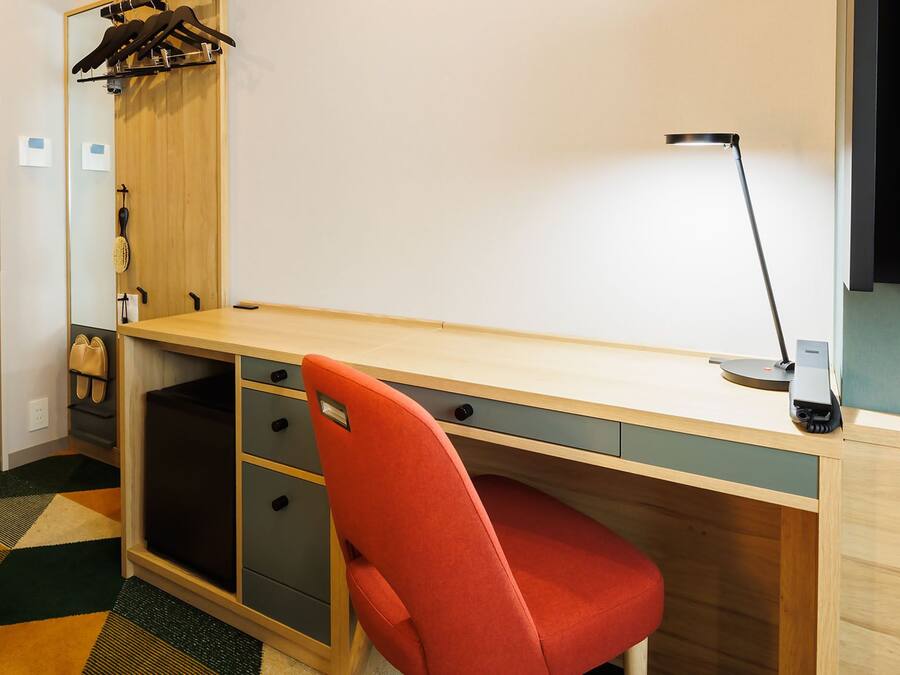 Habitación de hotel pequeña con un escritorio de madera, una silla roja, una lámpara de escritorio y un televisor de pantalla plana.