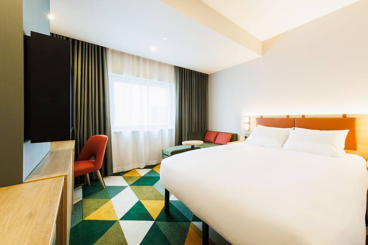 A hotel room with a large bed, a desk, a chair, a small table, and a TV.