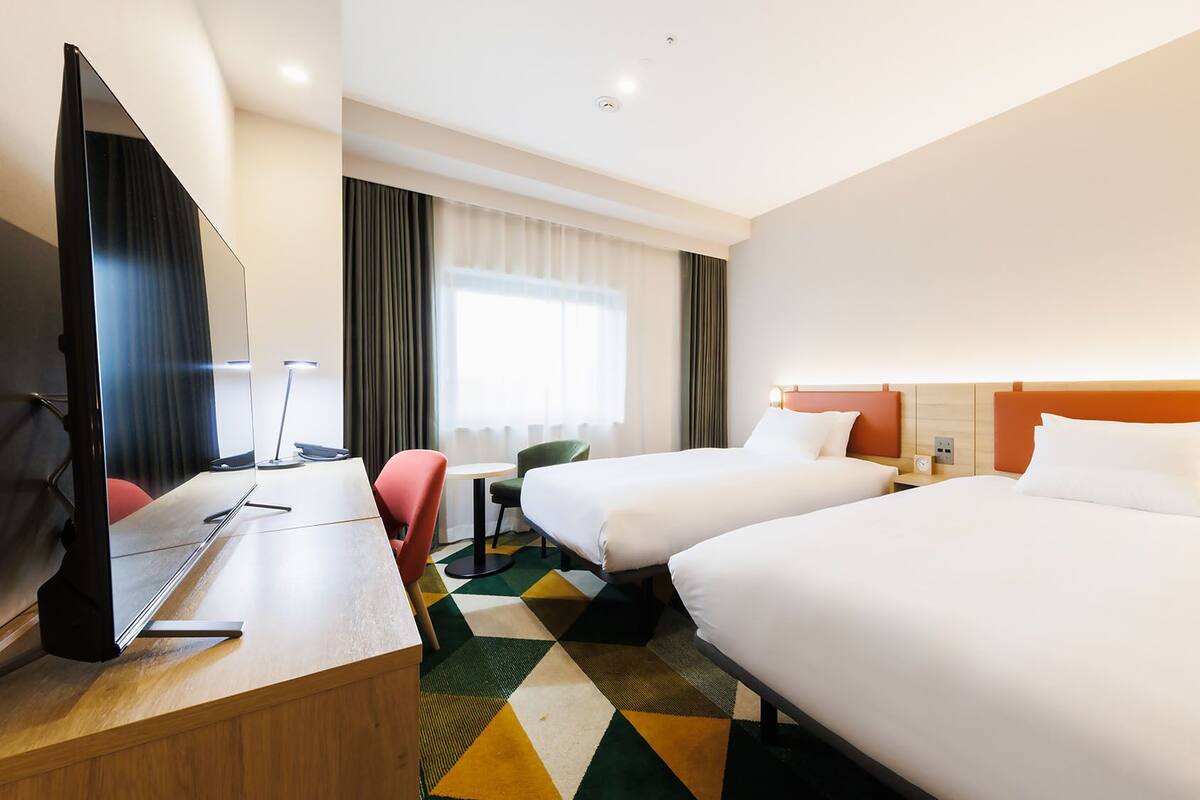 A hotel room with two beds, a desk with a computer, a TV, and a window with curtains.