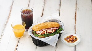 Un sándwich en una cesta, un vaso de zumo de naranja y un bol de granola sobre una mesa de madera blanca.