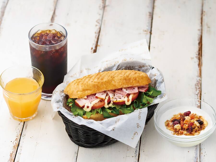 Ein Sandwich in einem Korb, ein Glas Orangensaft und eine Schüssel mit Granola auf einem weißen Holztisch.