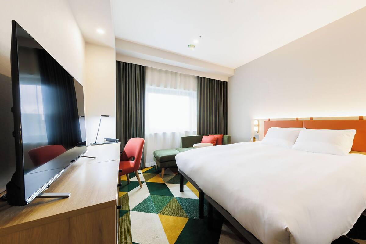 A hotel room with a bed, a desk with a computer, a TV, a chair, a sofa, a nightstand, and a window with curtains.