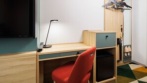 Habitación de hotel compacta con escritorio de madera, una silla roja, un refrigerador pequeño negro, televisor y un zapatero.
