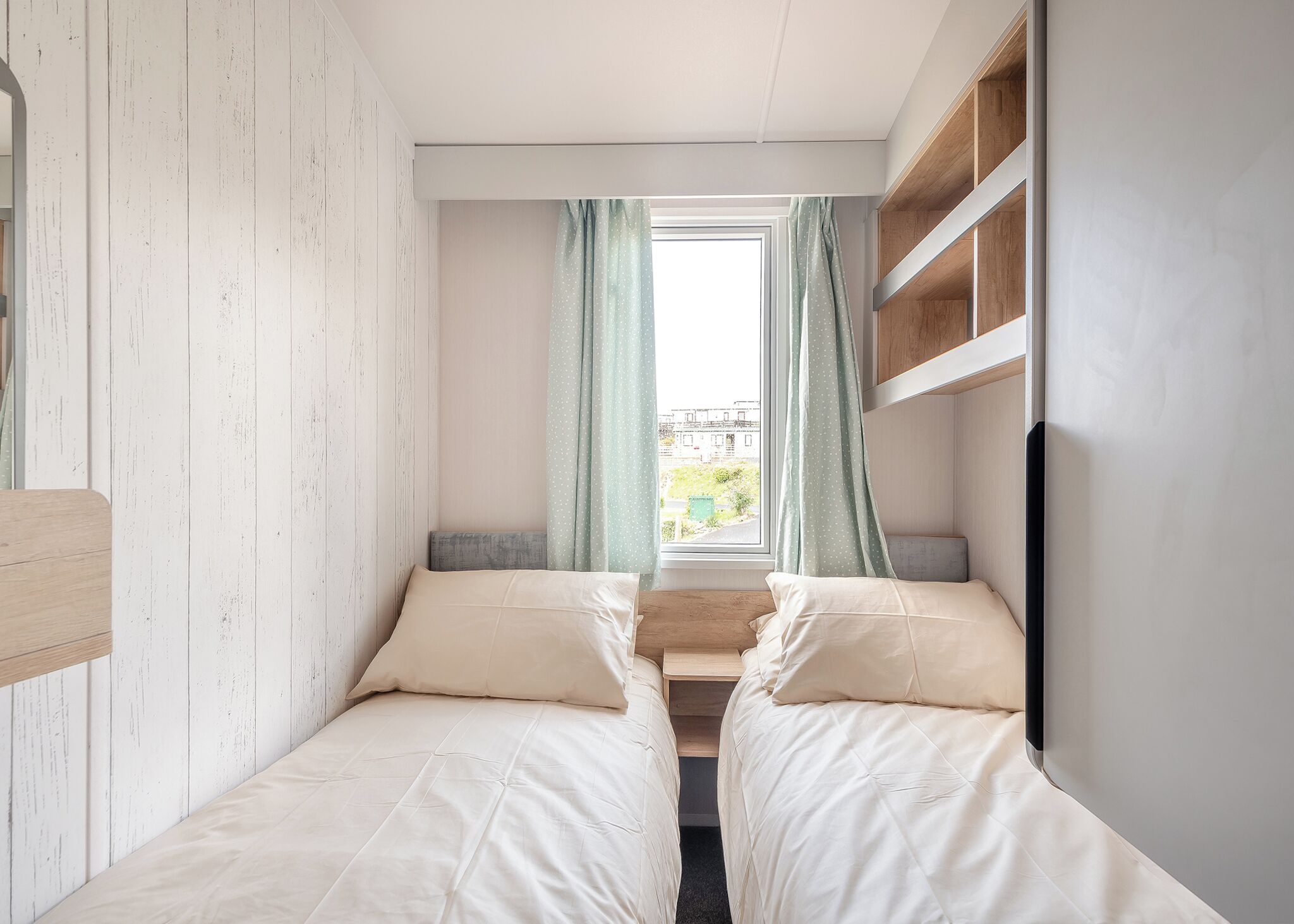 A small, modern room with two beds, a window with curtains, and a built-in shelf.