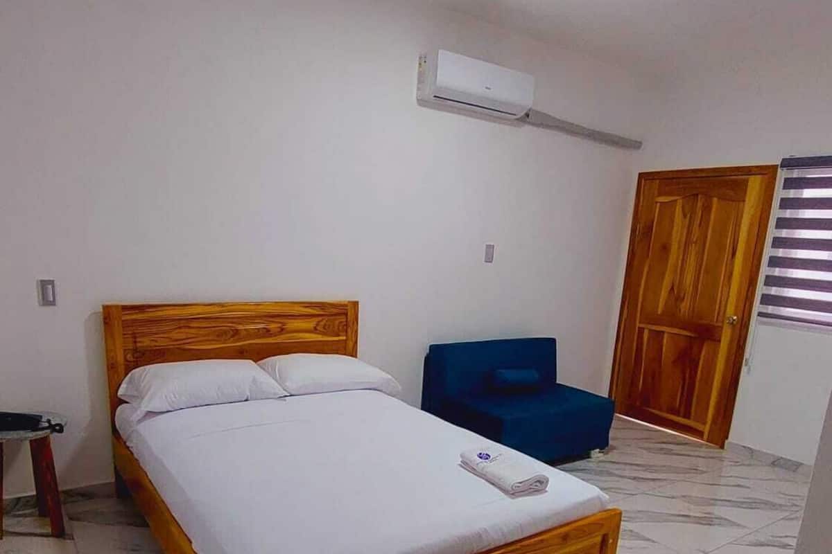 Un dormitorio con una cama de madera, una silla azul y un equipo de aire acondicionado en la pared.