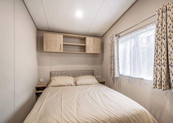 A bedroom with a bed, a nightstand, a built-in wooden cabinet, a window with patterned curtains, and a ceiling light.