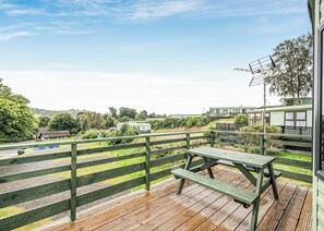 Una terraza de madera con una mesa de picnic, un área cercada y vistas de un edificio verde con ventanas y un balcón.