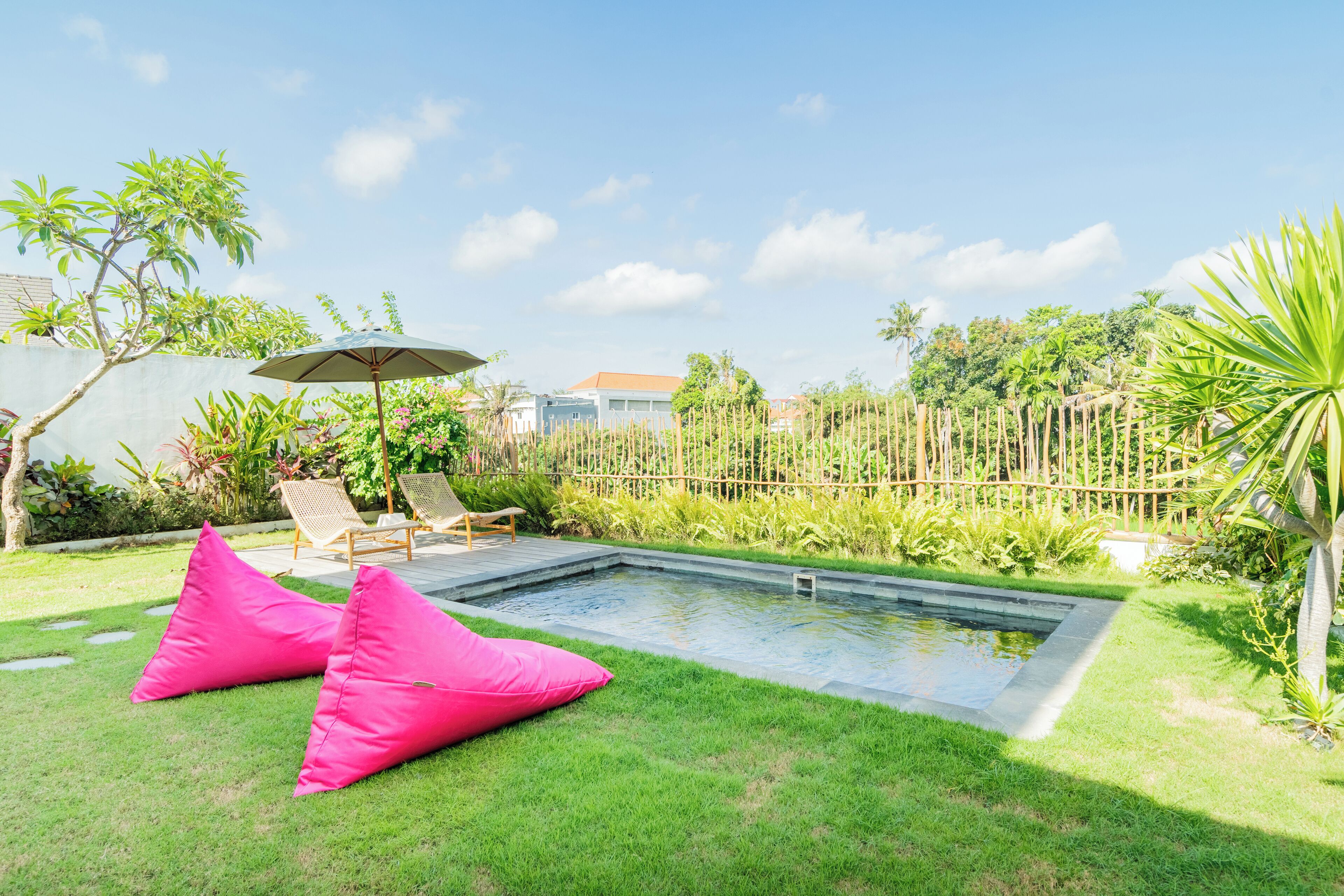 Un jardin avec une piscine, deux poufs roses, une clôture en bois et une variété de plantes.