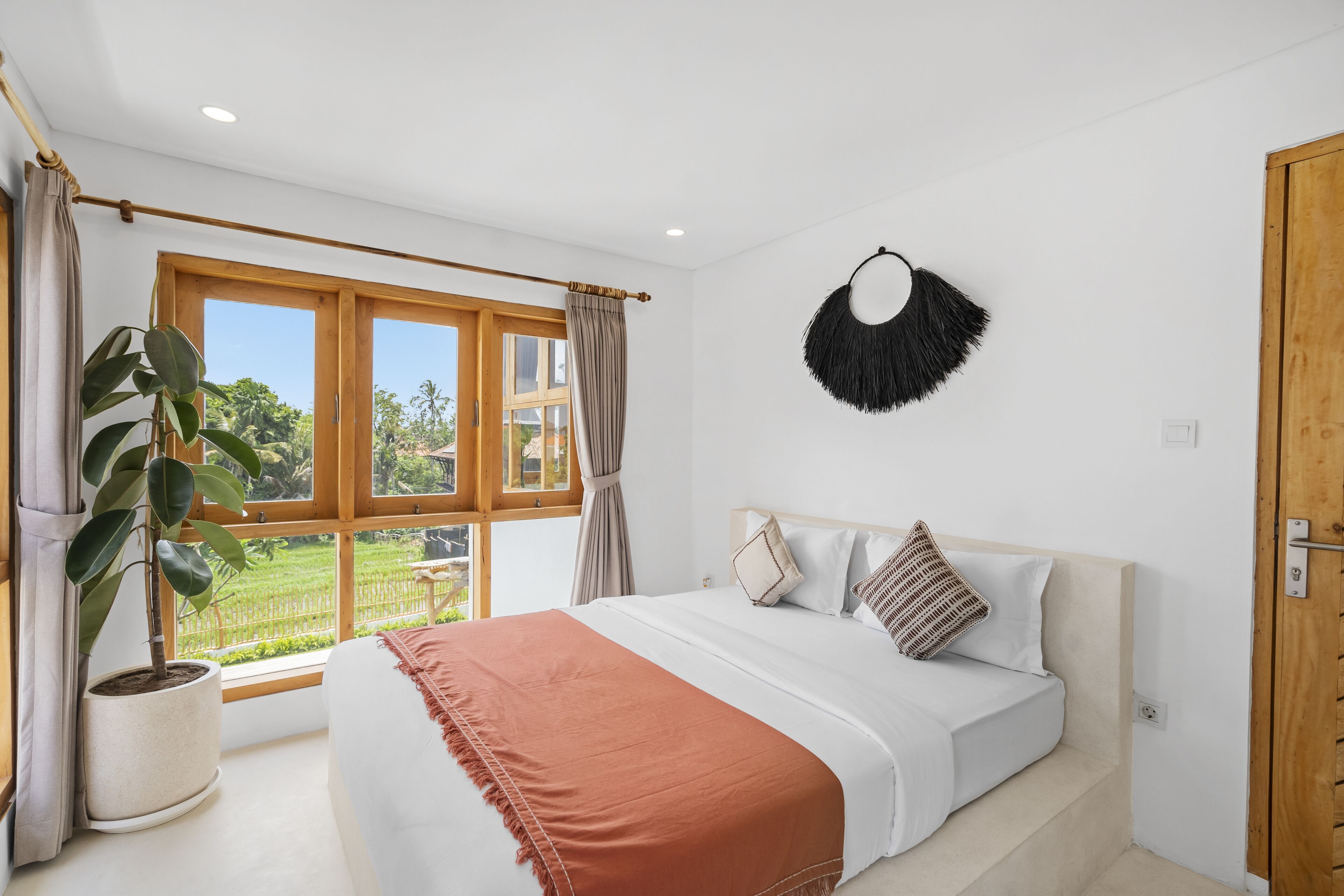 A bedroom with a large window, a potted plant, a bed with white bedding and a brown throw, and a wooden door.