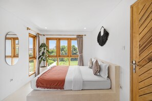 A bedroom with a bed, a window with curtains, a potted plant, and a wooden door.