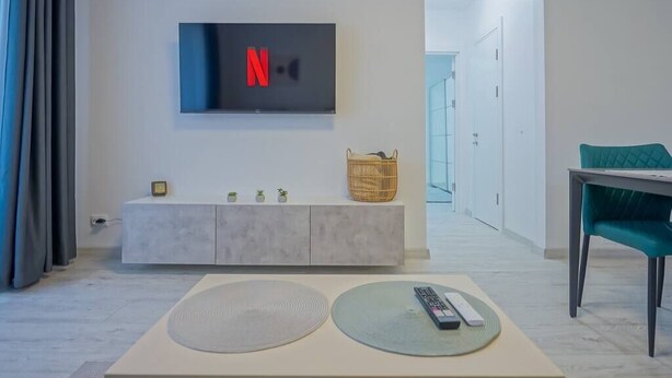 A modern living room with a TV mounted on the wall displaying the Netflix logo, a minimalist console table with a remote control, and a teal chair. - Mountain Hideaway w. Netflix and Mountain View (Brașov)