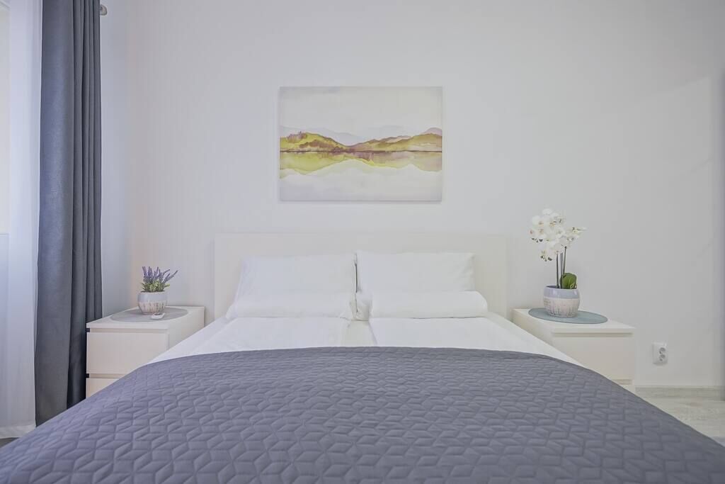 Ein Schlafzimmer mit einem großen Bett, weißer Bettwäsche, einem Landschaftsbild, zwei Nachttischen mit Blumentöpfen und einem Fenster mit Vorhängen.
