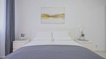 Ein Schlafzimmer mit einem großen Bett, weißer Bettwäsche, einem Landschaftsbild, zwei Nachttischen mit Blumentöpfen und einem Fenster mit Vorhängen.