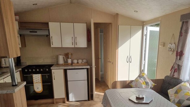 A compact kitchen with a stove, oven, and refrigerator, a dining area with a table and chairs, and a large window with curtains. - Robin hood rhyl, over the road from the beach (Rhyl)