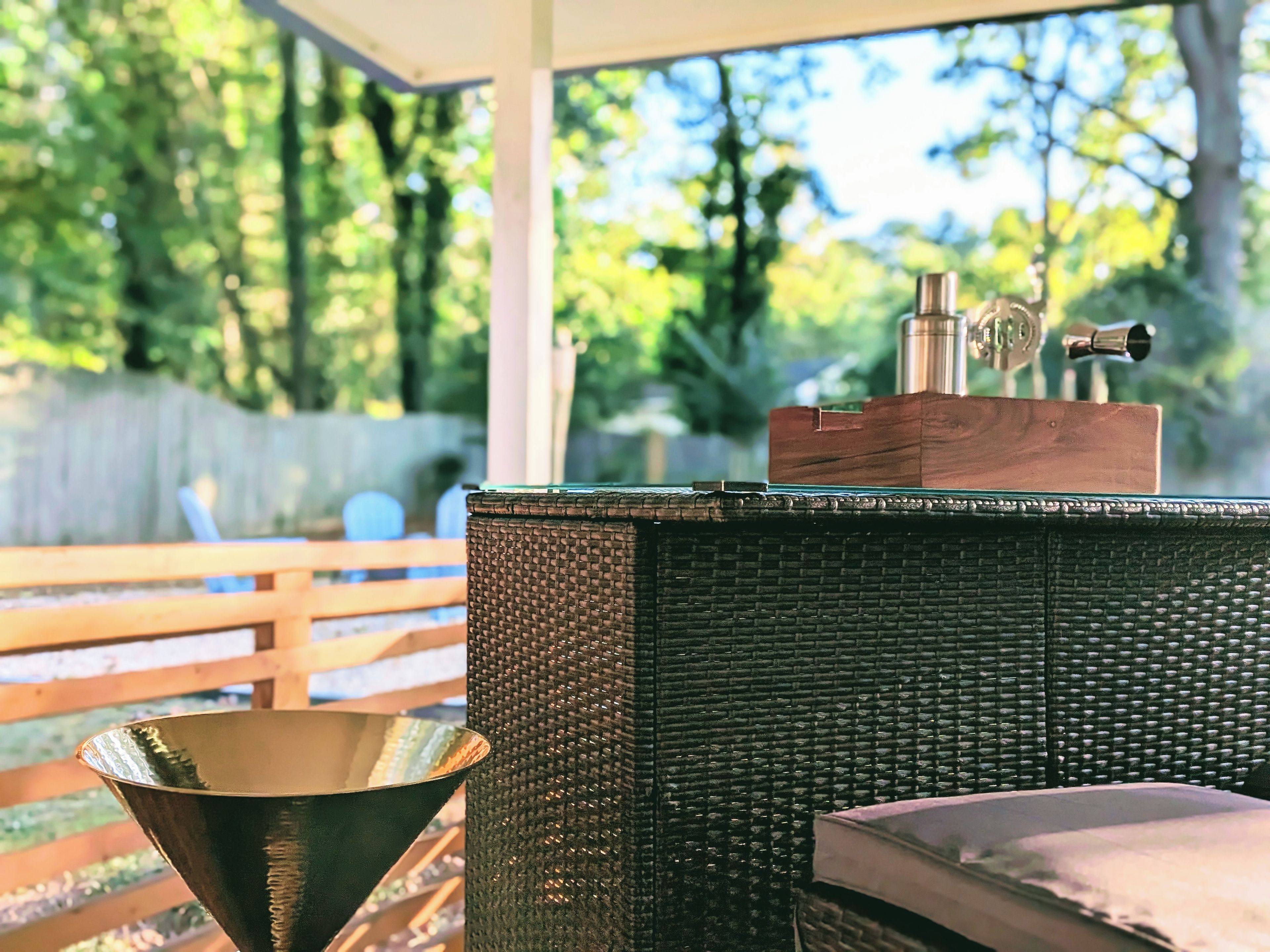 A covered outdoor seating area with a wicker table, a metal bowl, and a wooden box with a metal object on top.