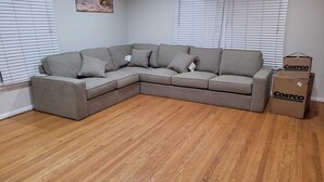 Un canapé d’angle gris, doté de plusieurs coussins, se trouve dans une pièce au plancher en bois et aux stores blancs.