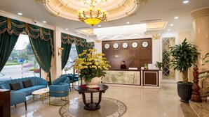 A hotel lobby with a reception desk, a chandelier, a large vase of flowers, and a seating area with blue sofas and a coffee table. - Móng Cái Place (Mong Cai)