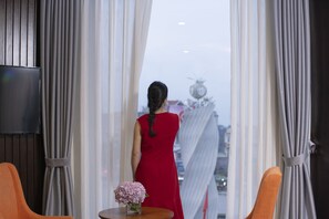 A room with a large window, a flat-screen TV, a small round table with a vase of flowers, and two orange chairs. - Móng Cái Place (Mong Cai)