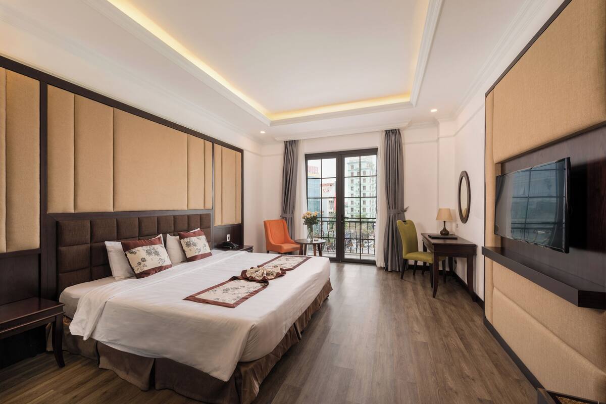 Habitación de hotel con una cama grande, televisión, escritorio y balcón con vistas a edificios.