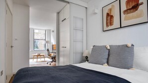 A bedroom with a large bed, a desk, and a chair. There is a window with a view of a brick building. - Clean Shoreditch 1BR w/ Open Plan, nr Market & Dining, by Blueground (London)
