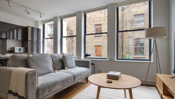 A modern living room with a grey sofa, a round wooden coffee table, and large windows overlooking a brick building. - Clean Shoreditch 1BR w/ Open Plan, nr Market & Dining, by Blueground (London)