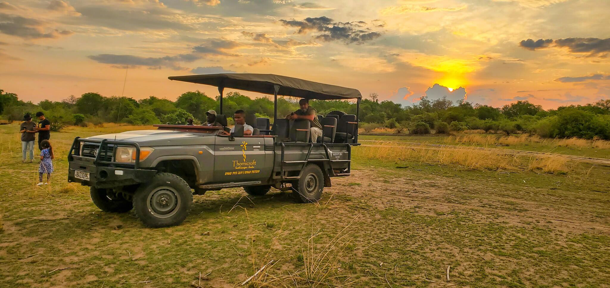Ein Safari-Fahrzeug mit Verdeck, geparkt in einem grasbewachsen Feld vor einem Sonnenuntergang.