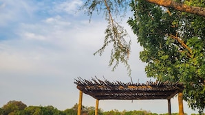Un pabellón con techo de paja y mobiliario de madera, rodeado por un sendero de ladrillo y una masa de agua.