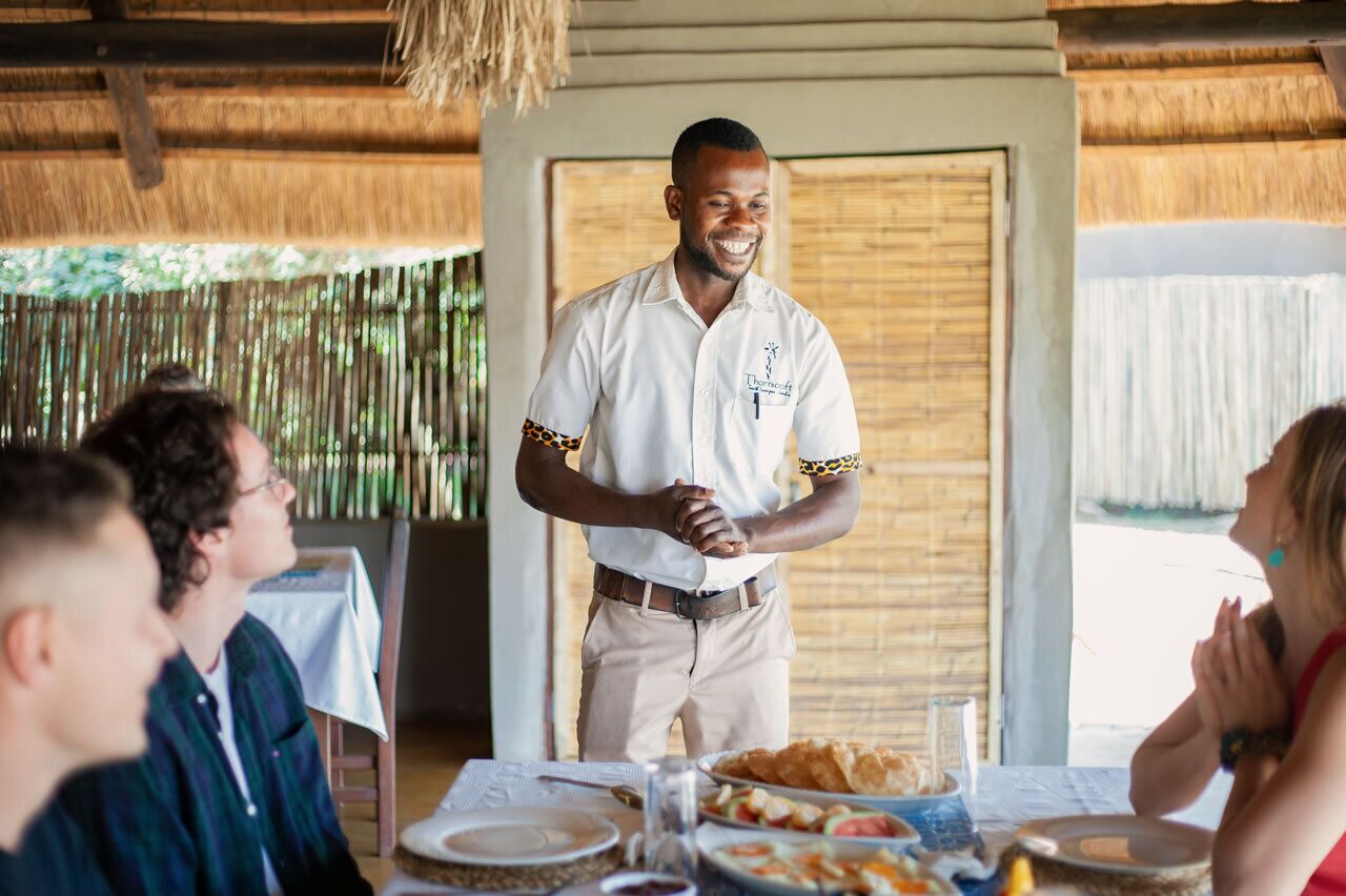 A person standing in front of a group of people at a table with food and drinks.