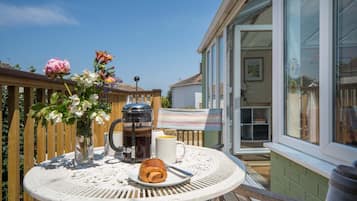 A round table with a coffee pot, mug, and croissant on a balcony with a wooden railing and a view of a house.