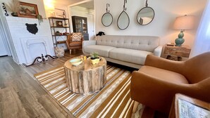 A cozy living room with a sofa, armchair, coffee table, and a fireplace. There are three wall-mounted mirrors and a lamp on a side table.