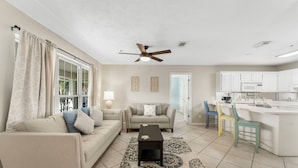 A living room with two sofas, a coffee table, a ceiling fan, and a kitchen area with white cabinets and a microwave.